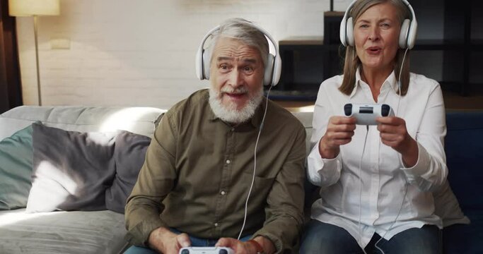 Attractive Caucasian Mature Couple in Headphones Playing Video Games at Home. Handsome Senior Man and Woman holding Joystick sitting on sofa while playing. PlayStation. Modern Technology. Older People