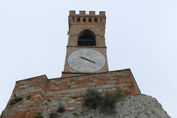 clock tower in Brisighella with mighty brick walls and the clock face at six hours on a hill that...