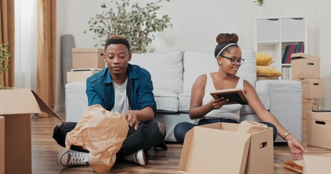Young people in love move into a new apartment, sit on the floor and pack their things into boxes, wrap vases, frames in paper, selling, renting a house