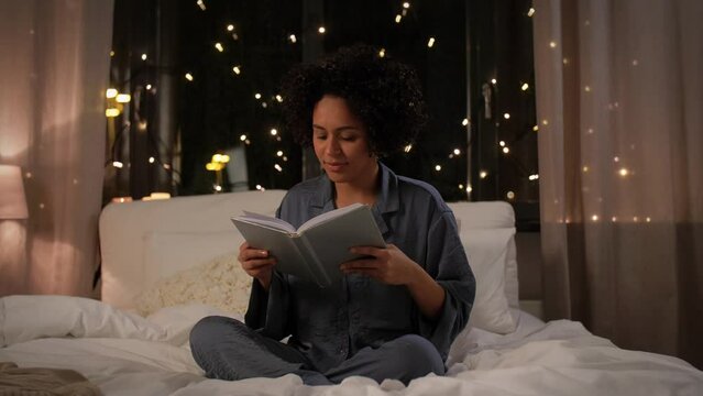 people, bedtime and rest concept - woman in pajamas reading book sitting in bed at night