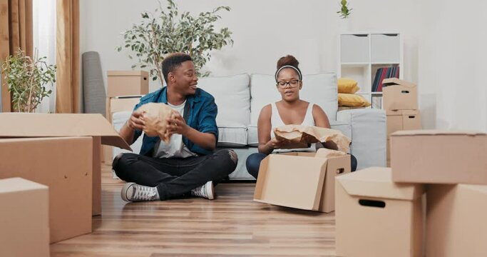 A married couple sits on the floor of their new apartment, unpacking boxes from the move, unrolling a paper photo frame, a keepsake vase, trinkets, smiling, spending time together