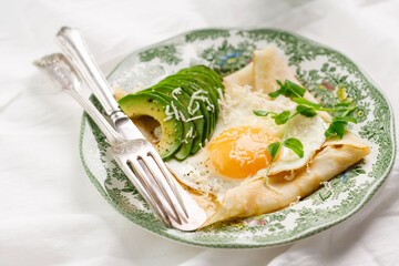 crepe with fried eggs, fresh avocado and cheese on a green vintage plate on a white textile background