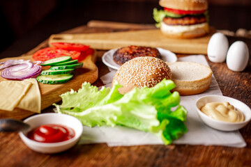 Cooking hamburger or cheeseburger. Different ingredients for a classic hamburger. Grilled meat, vegetables, greens, sauces near a sesame bun. Wooden background.