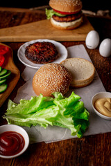 Cooking hamburger or cheeseburger. Different ingredients for a classic hamburger. Grilled meat, vegetables, greens, sauces near a sesame bun. Wooden background.