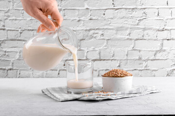 Vegan non diary buckwheat milk is pouring into a glass, near is buckwheat groats a bowl on a white...