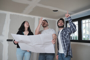 Latin male architect pointing at the ceiling and showing new apartment in construction blueprints to a young caucasian couple interested in buying.