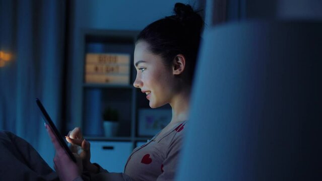technology, bedtime and people concept - happy smiling teenage girl tablet pc computer sitting in bed at home at night