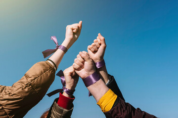 4 women's fists with a purple ribbon on the wrist with the blue sky in the background. Claim...