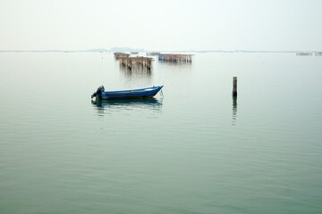 Barca azzurra immersa in atmosfera calma e misteriosa del mare piatto con sfondo allevamenti di vongole e cozze nel mare adriatico presso Chioggia Venezia Italia
