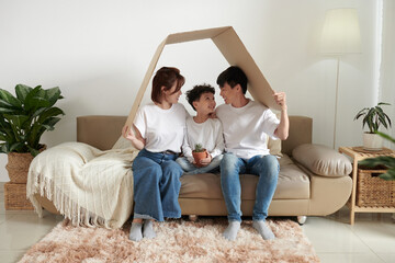 Happy parents and son sitting on sofa under cardboard roof, property insurance concept