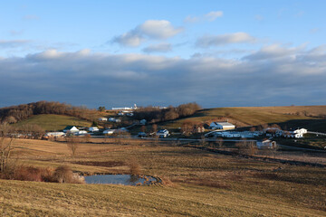 Amish Farms and Homesteads Surrounded by a Valley of Brown Fields and Bare Trees, Underneath a Partly Cloudy Blue Sky with a Small Pond in Foreground