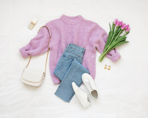 Lilac knitted sweater, blue jeans, white sneakers, bag and bouquet of tulips flowers lie on white background. Overhead view of woman's casual outfit. Trendy stylish women clothes. Flat lay, top view.