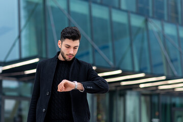 Young caucasian business man looking at his smart watch, business person checking time, modern financial skyscraper on back ground