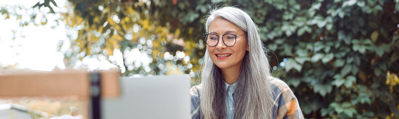 Happy mature Asian woman with glasses and strawberry dessert sits in front of laptop on table on outdoors cafe terrace on autumn day