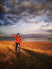A beautiful girl in a bright orange jacket rides a bicycle across the field at sunset, against the backdrop of beautiful clouds.