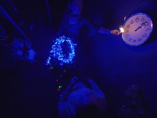 The family fast and fanny decorates the Christmas tree. Top view. Time on the clock is 23.45 on wall. Happy dad, mom and daughter are running around tree, hanging colored balls, garlands.