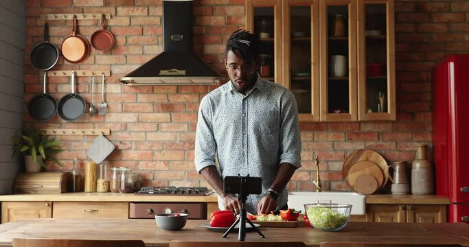 Charismatic young Black man food blogger hipster shoot culinary tutorial at home kitchen on smartphone. Enthusiastic millennial guy master chef record vlog webcast live teach to cook fresh vegan salad