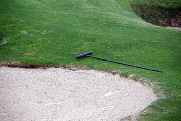 Partial view of a golf course sand trap bunker and a rake next to it on the grass - Powered by Adobe