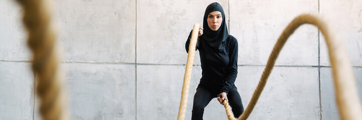 Young muslim woman in hijab working out with battle ropes indoors