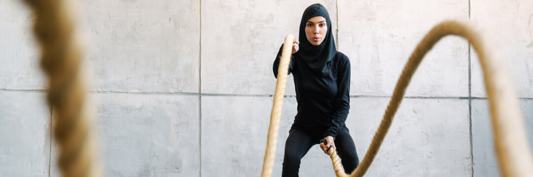 Young Muslim Woman In Hijab Working Out With Battle Ropes Indoors