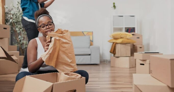 A married couple moves into new apartment woman sits on floor unpacking cardboard boxes of belongings unrolls picture frame from paper man puts pillows on couch and sits down relaxing