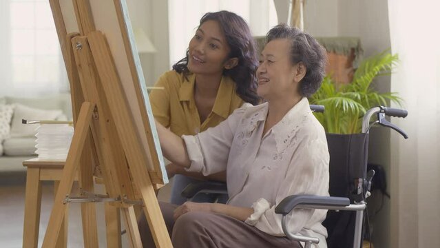 Female teacher teaching senior woman to paint picture at living room. Senior woman sitting wheelchair while paint picture. Healthcare, Lifestyle and Hobby concept.