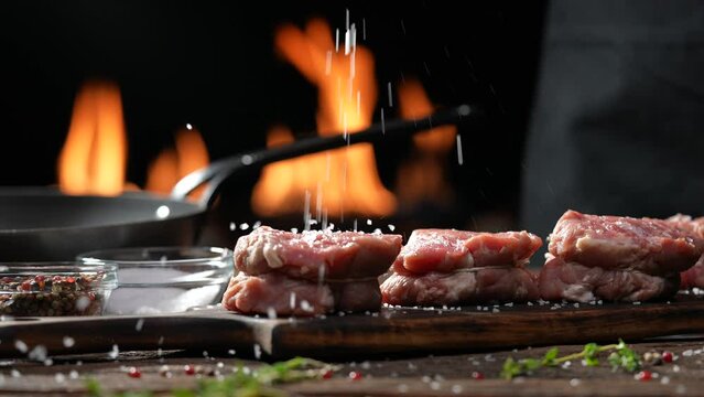 Hand sprinkling salt and seasoning on raw tenderloin steak meat beef on wooden chopping board on a wooden table prepared for cooking with flames in the background. slow motion