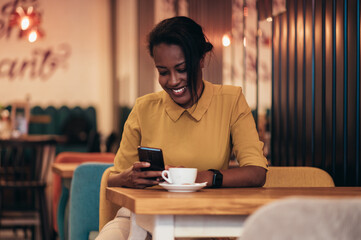 African american woman using a smartphone and drinking coffee while sitting in a cafe