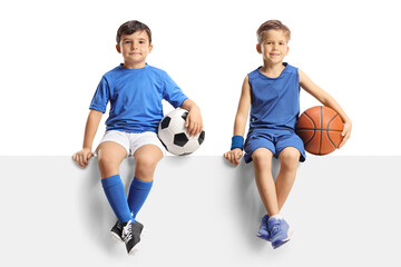 Two boys in sport jersey sitting on a panel with a football and a basketball