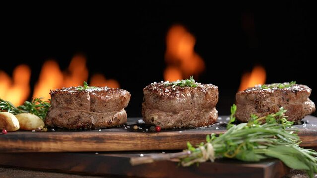 Cooked juicy steak meat beef with hand sprinking seasoning on top on wooden chopping board with flames in the background. slow motion