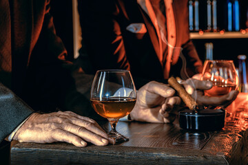 Man's hands with a cigar, elegant glass of brandy on the bar counter. Alcoholic drinks, cognac,...