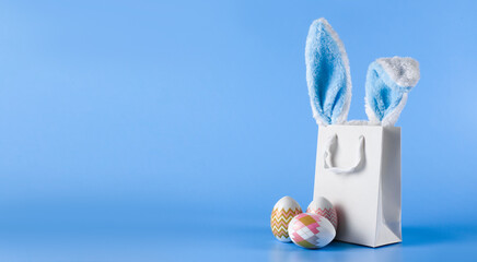 Wide banner on a blue background. The concept of online shopping for Easter, purchases and sale for the holiday. Gift bag with bunny ears and Easter eggs