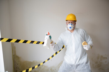 Man with protective mask, protective suit and yellow safety helmet tries to remove mold on wall...