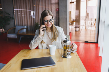 Successful young woman solving business problems during a telephone conversation, sitting in a modern cafe and working with a laptop. Cute interior is enjoying his free time in the cafe.