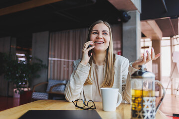 Young attractive female manager is talking on a mobile phone and smiling while sitting alone near a large window in a cafe in her free time and working on a computer. Happy woman is resting in a cafe.