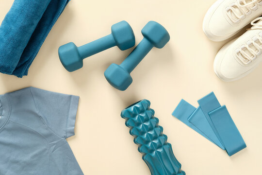 Fitness equipment for workout on beige table. Dumbbells, fitness tape, roller, female t-shirt and sneakers.
