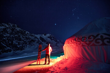 A man with a snowboard standing near a yurt at night in winter in the mountains