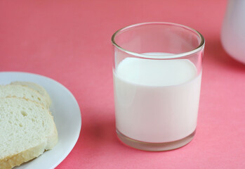 A glass of milk stands on a pink table, next to it lies bread in a white plate. Behind is a milkman