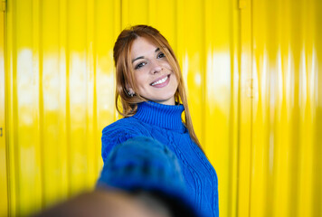 Selfie looking at camera of a redhead girl with blue sweater and yellow background smiling.