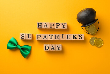 Top view photo of wooden cubes labeled happy st patricks day green bow-tie and pot with golden coins on isolated yellow background