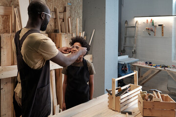 Portrait of black father and son working together in garage workshop, focus on smiling boy wearing...