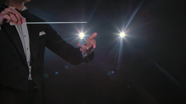 Moving hands of an orchestra conductor directing musicians with movement of baton on black background with lights. Conducting, directing classical musical performance with visible gestures. Close up.