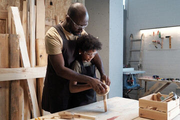 Waist up portrait of caring African-American father teaching son in carpentry workshop, copy space