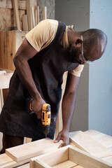 Vertical portrait of black carpenter using power drill while building wooden furniture in workshop