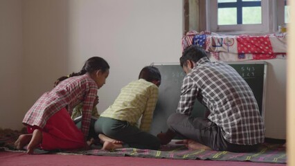 A young Indian rural school male teacher is writing on a blackboard while teaching a small group of children in a  classroom in a countryside or village area. Concept of education and learning