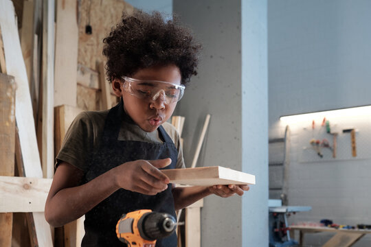 Portrait of cute black boy blowing sawdust off piece of wood in carpentry workshop, copy space