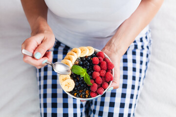 The girl eats a healthy breakfast of muesli with fresh fruits and berries while sitting on the bed...