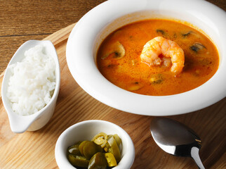 Tom Yam on a light wooden background. Serving soup in a white plate in a restaurant. Thai traditional dish with rice and shrimp. Delicious lunch. Close up, Copy space. Delicious soup with a spoon