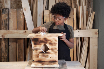 Front view portrait of teenage African-American boy building wooden birdhouse in workshop, copy...