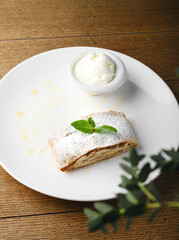 Apple strudel with cinnamon and a scoop of ice cream. Traditional Austrian dessert on a white plate on a wooden background. Close up. Copy space. Delicious apple pie for kids.
